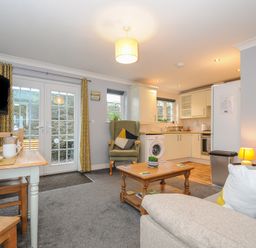 An open living room and kitchen with a sofa, armchair, coffee table, dining table set near French doors and kitchen cabinets at 1 Bethany Court in Bodmin