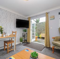 A living room with a small dining table set for breakfast a television mounted on the wall and French doors leading to an outdoor area at 1 Bethany Court in Bodmin