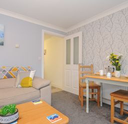 A living room with a beige sofa pillow and a wooden table with plants and coasters and a dining table set with flowers and breakfast at 1 Bethany Court in Bodmin