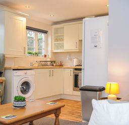 A kitchen area with white cabinets, a washing machine, a refrigerator, a trash bin, a wooden coffee table with coasters and a small plant, an armchair with cushions, and a side lamp at 1 Bethany Court in Bodmin