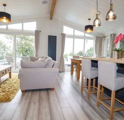 A living room and kitchen area with light gray sofas wooden tables and chairs and large windows at Mordros Lodge in St Day