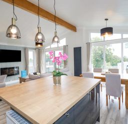An open plan living and dining area with a wooden island table and pink flowers a dining table with chairs large windows and a mounted television at Mordros Lodge in St Day