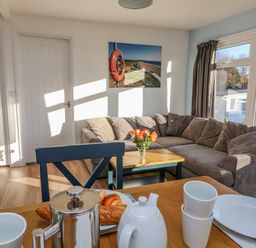 A living room with a grey striped corner sofa a wooden coffee table with flowers and a dining table set with white cups plates and a stainless steel coffee pot at Beachcomber Norton Park near Dartmouth