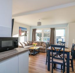 A dining area with a wooden table and four chairs next to a living room with a corner sofa and windows at Beachcomber in Norton Park near Dartmouth