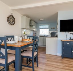 A dining area with a wooden table and four chairs next to a kitchen and a wall-mounted TV at Beachcomber Norton Park near Dartmouth