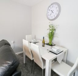 A dining area with a white table and four white chairs a bottle of wine and plates on the table a black leather couch and a wall clock at 17 Ffordd Gwenllian in Llanfairpwllgwyngyll