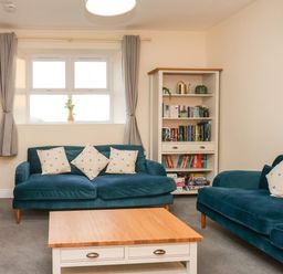 A living room with two blue sofas a wooden coffee table a bookshelf and a window with curtains at Garden Cottage in Alnmouth