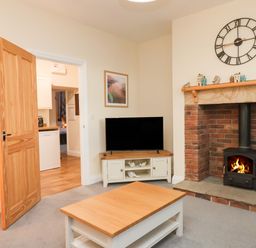 A living room with a wooden coffee table a TV on a stand an open wooden door and a brick fireplace with a fire burning at Garden Cottage in Alnmouth