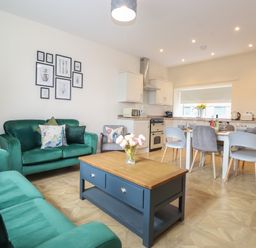 A combined living room and kitchen with green sofas a wooden coffee table dining table with chairs and kitchen appliances at Pond Cottage in Alnmouth