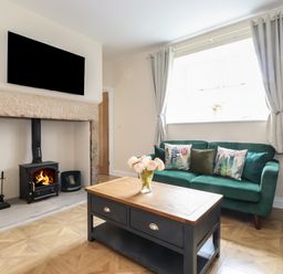 A living room with a green sofa, coffee table with flowers, wood burning stove, and wall-mounted television at Pond Cottage in Alnmouth