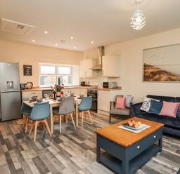 An open plan kitchen and living room with a dining table set for six and blue and grey chairs at Lonnon Cottage in Alnmouth