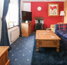 A living room with blue patterned carpet a wooden coffee table a television on a wooden stand a red accent wall with a clock and flower painting at Bod Feurig Penygroes