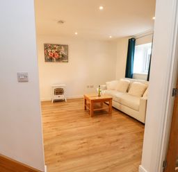 A living room with a white sofa with cushions a wooden coffee table with a bottle and glasses a small heater and a painting on the wall at Stable Cottage in Hallbankgate