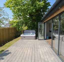 An outdoor deck area with a hot tub next to a glass sliding door showing a living room with a sofa at The Nest Brython in Tenby