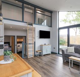 A living room with a gray sofa a small wooden table a wall-mounted TV and large windows at Cornerstones Brython in Tenby