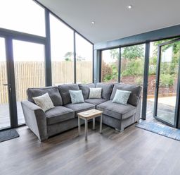 A corner seating area with a grey sectional sofa and small wooden table in a room with large glass windows and doors at Cornerstones Brython in Tenby