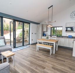 A living area with a gray sofa and pillow a small wooden table a wooden dining table with chairs and bench kitchen cabinets and appliances large glass doors opening to a garden at Cornerstones Brython in Tenby