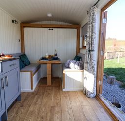 A small dining area with built-in benches and a wooden table next to a kitchen with cabinets and an open door showing an outdoor grass area at Cariad Bach in Gwaenysgor near Prestatyn