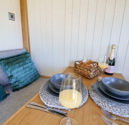 A dining table set for two with gray dinnerware glasses of orange juice a bottle of sparkling wine and a basket of bread with a cushioned bench seat at Cariad Bach in Gwaenysgor near Prestatyn
