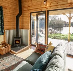 An interior with a wood burning stove a green sofa with cushions a basket with firewood and large glass doors overlooking a pond at Kingfisher in Dunchurch