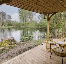 A covered wooden patio with two chairs and a table overlooking a pond surrounded by trees at Dragonfly in Dunchurch