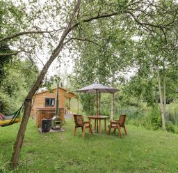 An outdoor area with wooden chairs around a table and umbrella a hammock and a small wooden cabin near a body of water at Dragonfly in Dunchurch
