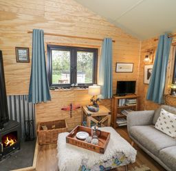 A living room with a wood stove a gray couch blue curtains and a tray on a ottoman at Dragonfly in Dunchurch