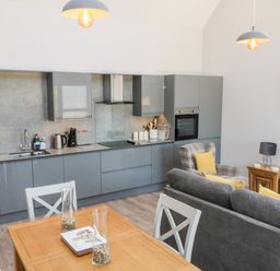 A kitchen with grey cabinets and appliances next to a dining table with white chairs and a living area with grey sofa and plaid armchair at Willow Cottage in Combe Martin