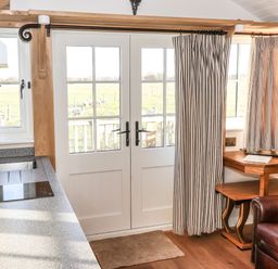 A kitchen area with a sink and stove next to glass doors with curtains and a corner wooden table by a leather armchair at The Egyptologist in Bolton Percy near Tadcaster