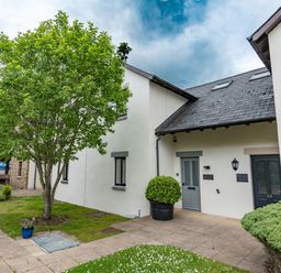 Exterior view of white buildings with grey doors and windows a tree and bushes on a paved walkway at Whisk Away Cottage in Greystoke near Troutbeck Near Penrith