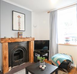 A living room with a fireplace wooden mantel a black TV on a stand a window with gray curtains an ottoman with a colorful blanket and a black coffee table with plants and candles at 5 Thomas Street in Llanberis