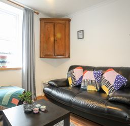 A living room with a black leather sofa with three colorful pillows a small black coffee table with a plant and candles a wooden corner cabinet and a window with grey curtains at 5 Thomas Street in Llanberis