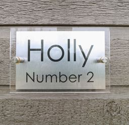 A metal and acrylic house sign on a textured wooden wall with the text Holly Number 2