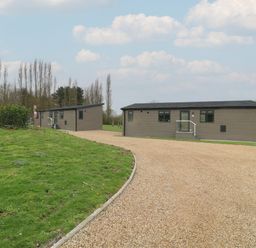 Two single-story grey buildings with black roofs on a gravel driveway next to a grassy area at Cedar Lodge in Newark-On-Trent