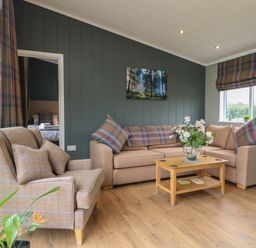 A living room with a beige sofa and armchair with plaid cushions a wooden coffee table with a flower vase and a framed forest picture on the wall at Cedar Lodge in Newark-On-Trent