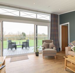 A living room with armchair sofa coffee table and sliding glass doors leading to a balcony with chairs at Cedar Lodge in Newark-On-Trent