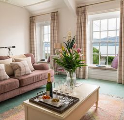A living room with a pink sofa and pillows a white coffee table with a flower vase a tray with drinks and snacks and windows with views of water at Tros Yr Afon in Beaumaris