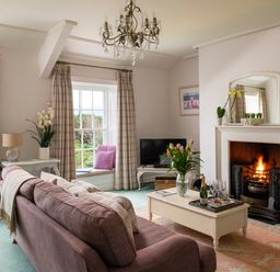 A living room with a purple sofa a coffee table with flowers and a bottle of wine a fireplace with a fire and large windows with curtains at Tros Yr Afon in Beaumaris