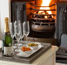 A tray with a bottle of champagne glasses and bowls of snacks near a lit fireplace at Tros Yr Afon in Beaumaris