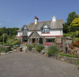 A two-story house with stone and white walls surrounded by greenery and a paved driveway at Storrs Lodge in Bowness-On-Windermere