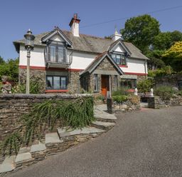 A stone and white house with a sloped roof a small balcony a wooden door and stone steps at Storrs Lodge in Bowness-On-Windermere