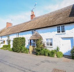 A long white thatched roof cottage with windows and a door surrounded by bushes and greenery at Lime Cottage in Burton Bradstock
