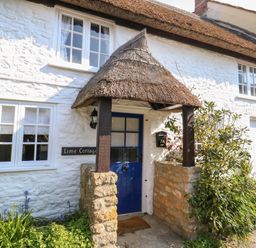 The entrance of a white stone cottage with a blue door and thatched porch at Lime Cottage in Burton Bradstock