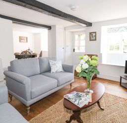 A living room with grey sofas a wooden coffee table with flowers in a vase and magazines a dining area visible through an open doorway at Lime Cottage in Burton Bradstock