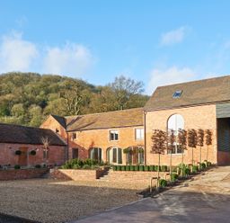 A brick building with multiple windows and a clock tower on the roof surrounded by gravel and small trees at Fairoaks Barns in Hollybush near Ledbury