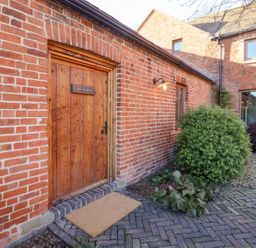 An exterior view of a red brick building with a wooden door labeled The Stables bushes and plant beds nearby at Fairoaks Barns in Hollybush near Ledbury