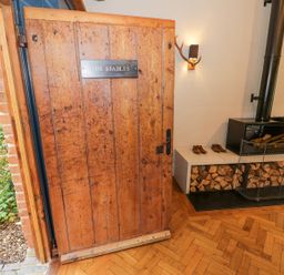 A wooden door with a sign that reads The Stables opening into a room with a fireplace with wood storage underneath and shoes on a bench at Fairoaks Barns Hollybush near Ledbury