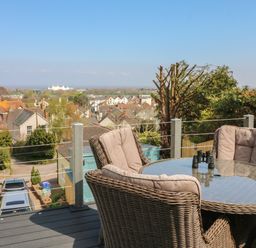 An outdoor seating area with a glass table and wicker chairs overlooking houses and trees at The Outlook in Minehead