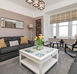 A living room with a dark gray sofa, armchairs, a white coffee table with yellow flowers, a floor lamp, window with plaid blind and small side tables at The Outlook in Minehead