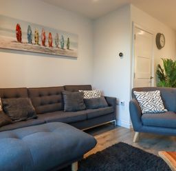 A living room with a gray sofa armchair ottoman pillows wall art clock and plant at Chy Fistral in Newquay